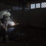 Operaio metalmeccanico al lavoro in un'industria, con attrezzature e macchinari in background.